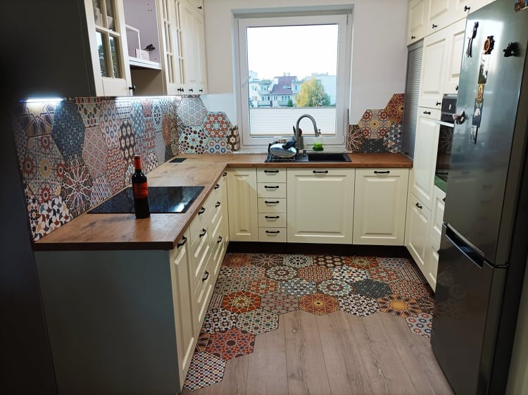 Wykończona kuchnia z białymi szafkami, drewnianym blatem i heksagonalnymi płytkami z wzorami arabesk na podłodze i ścianie, butelka wina na blacie.