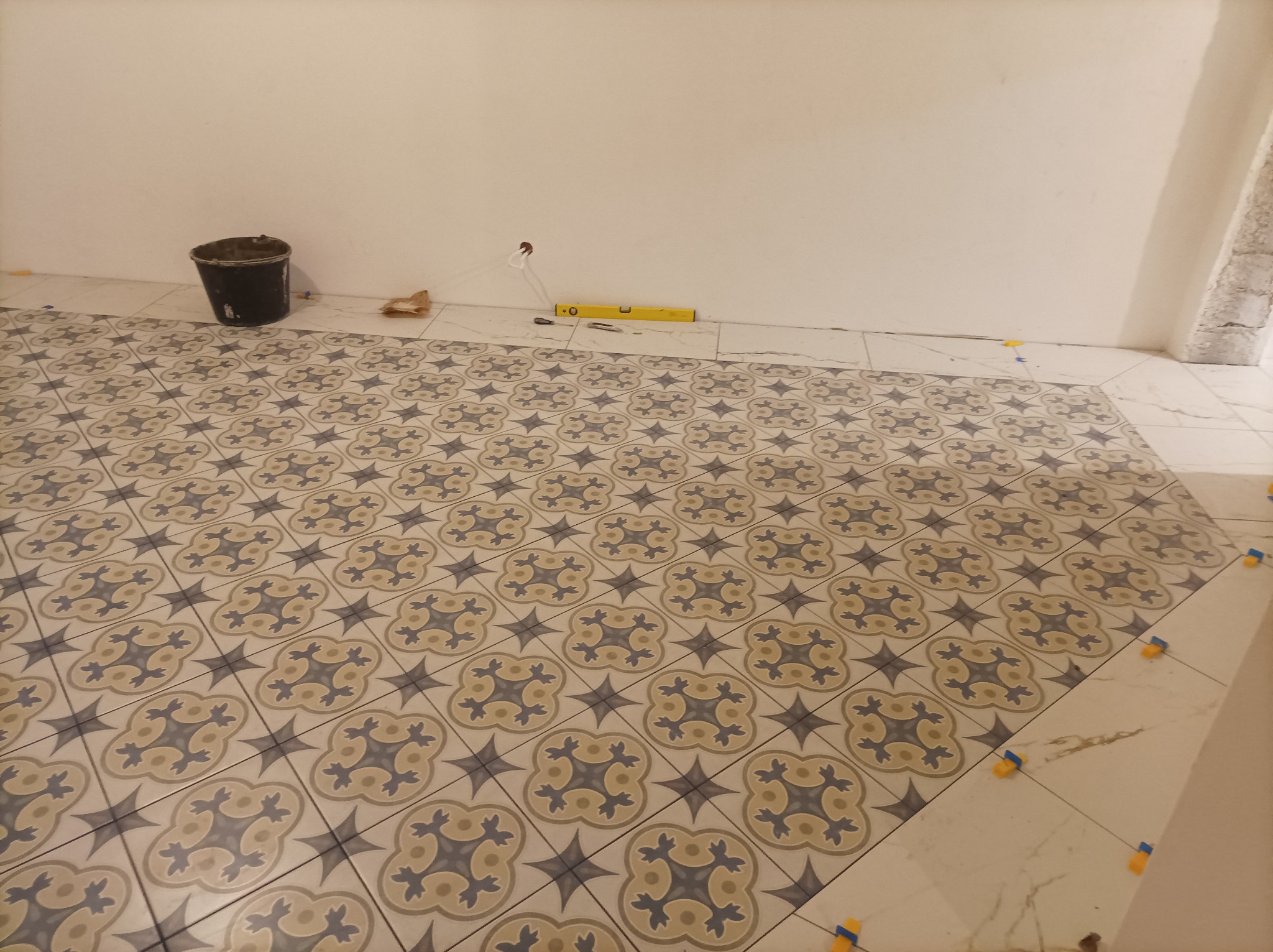 Układanie wzorzystych płytek podłogowych w pomieszczeniu, widoczny poziomica, wiadro i krzyżyki dystansowe.