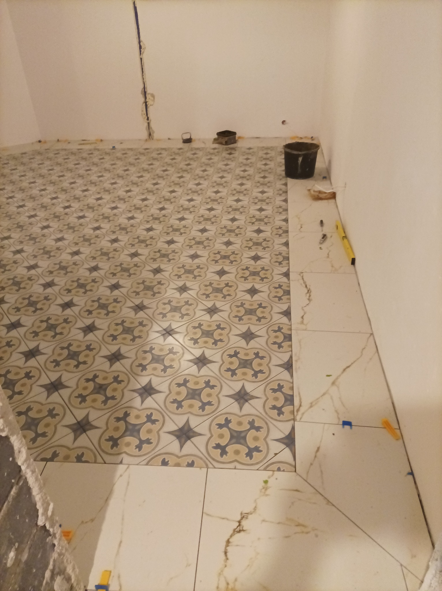 Układanie płytek podłogowych: wzorzyste płytki na podłodze, obok płytki imitujące marmur, poziomica i wiaderko widoczne w tle, etap prac.