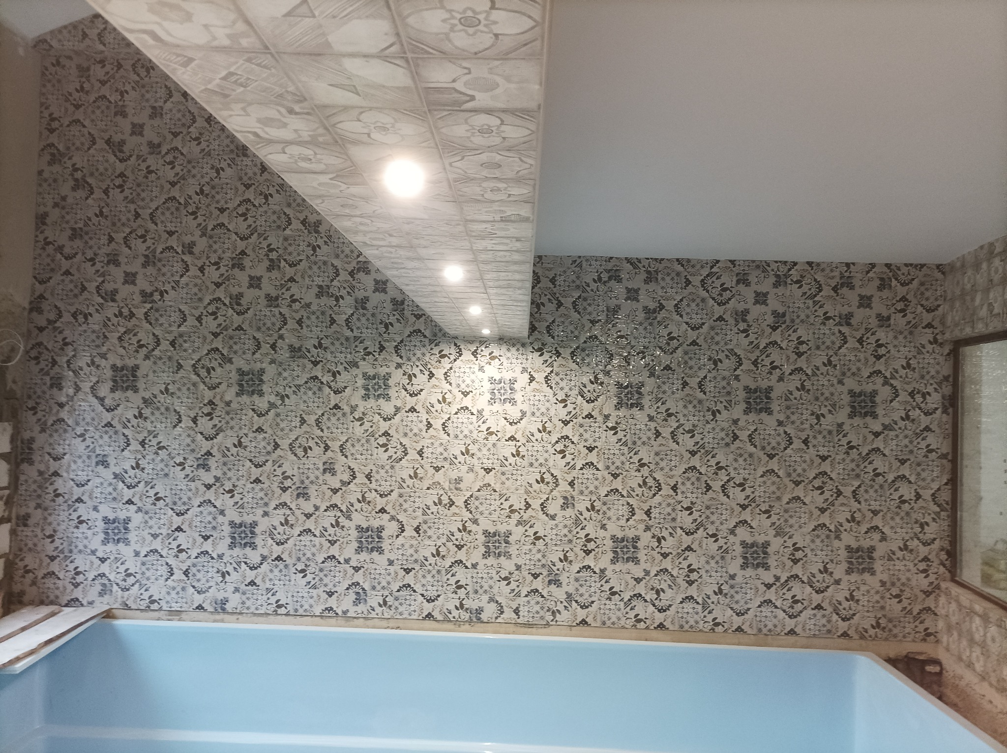 Łazienka w trakcie remontu: niebieska wanna, ściany i sufit wyłożone wzorzystymi płytkami, widoczne oświetlenie sufitowe.