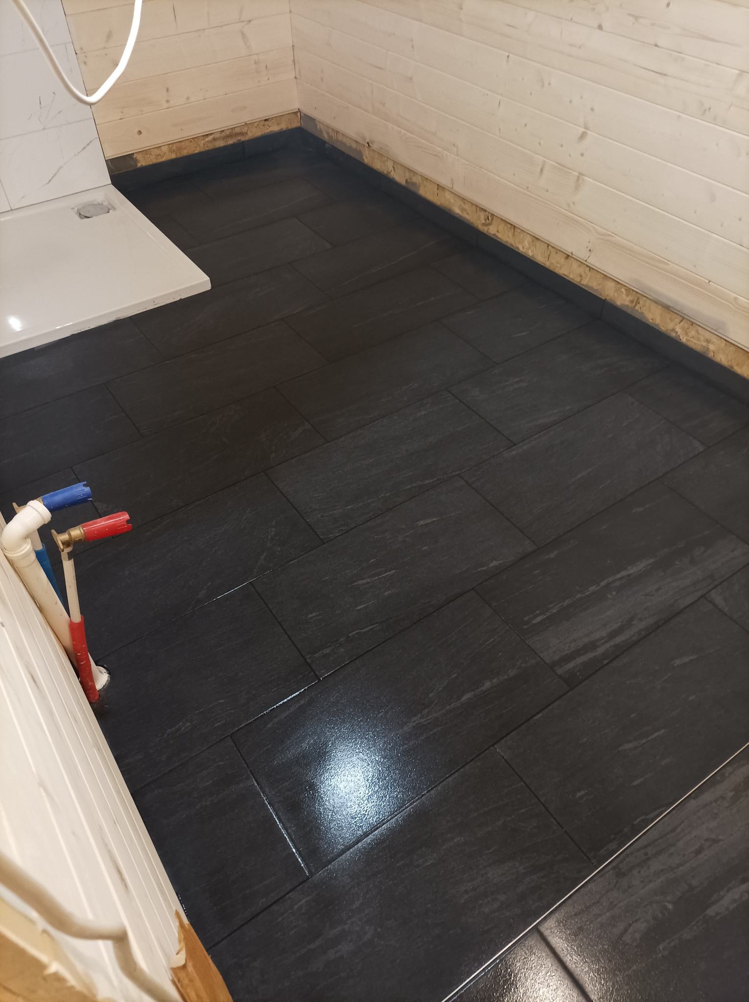 Ułożona, ciemnoszara podłoga z płytek imitujących kamień w łazience, widoczne fragmenty instalacji wodnej z czerwonym i niebieskim zaworem, ściany w jasnej boazerii.