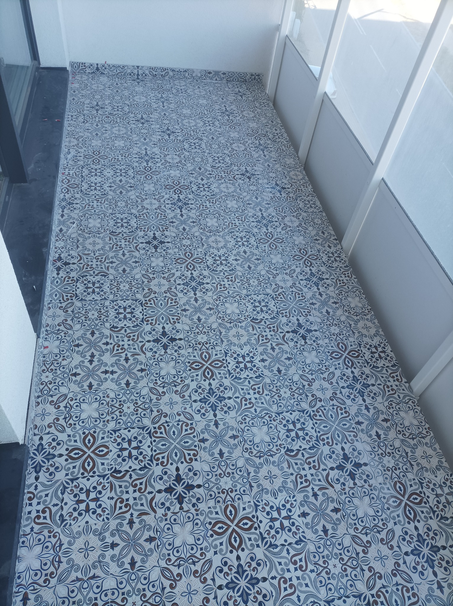 Wzorzyste, ceramiczne płytki podłogowe w odcieniach błękitu, szarości i brązu na balkonie, widoczne białe balustrady i fragment okna.