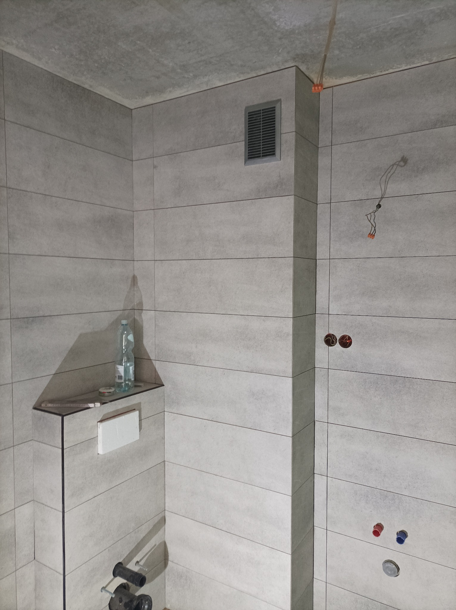 Szara łazienka w trakcie wykańczania: widoczne płytki imitujące beton, wystające kable, rury i niedokończona instalacja podtynkowa. Na półce butelka wody i narzędzia.