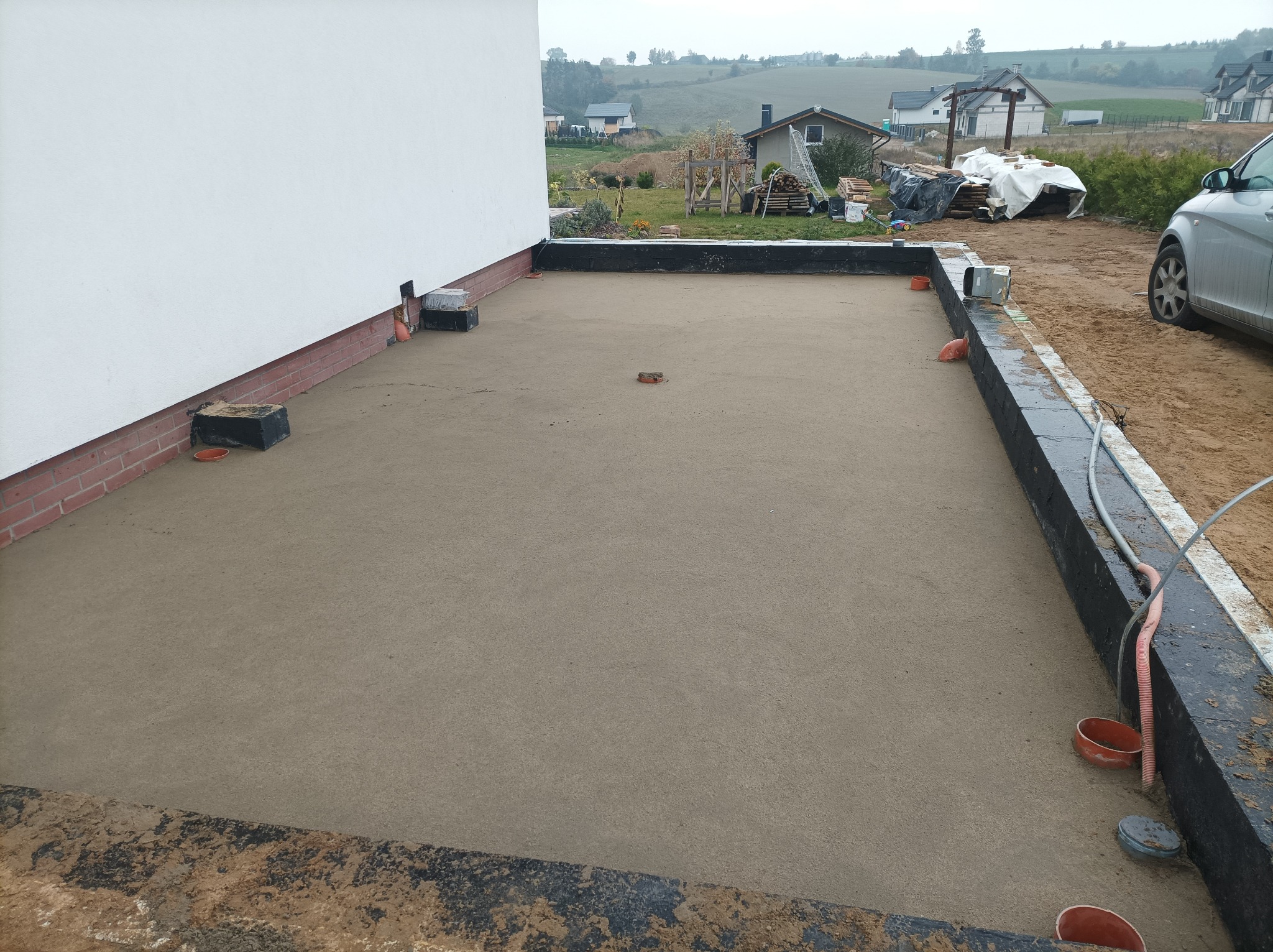 Przygotowany podkład pod taras z widocznymi elementami instalacji odprowadzającej wodę i fragmentem elewacji budynku jednorodzinnego w tle.