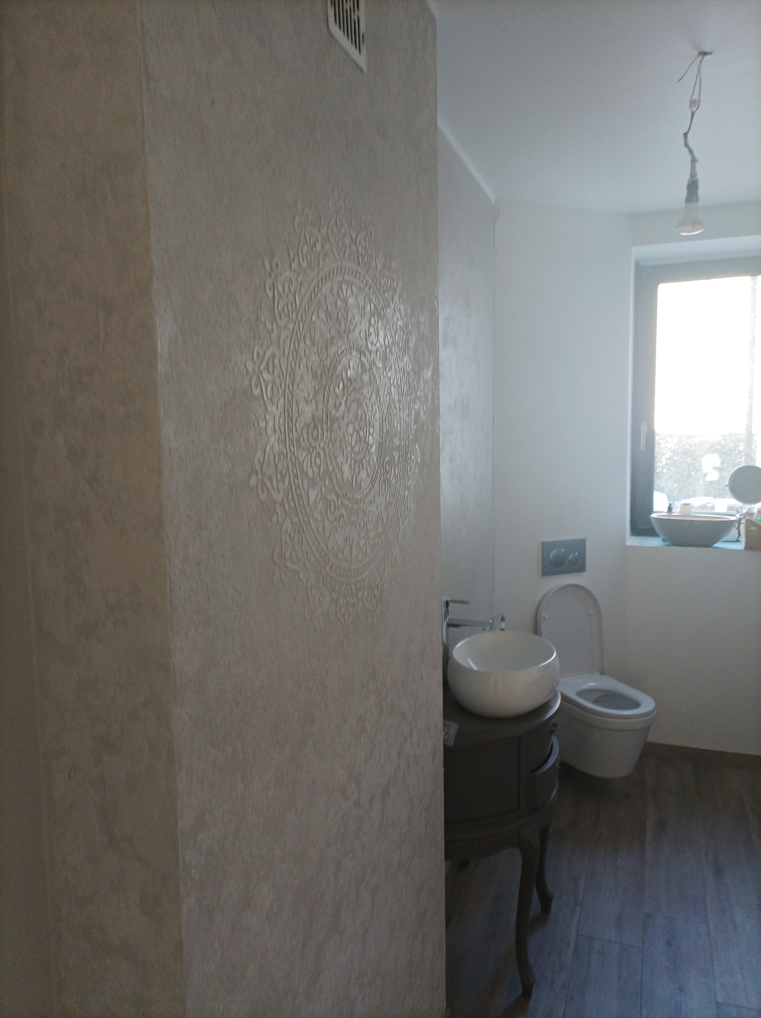 Wykończona łazienka z dekoracyjną ścianą w odcieniach beżu z ornamentem, okrągła umywalka na ciemnej szafce, podwieszana toaleta i drewnopodobna podłoga.