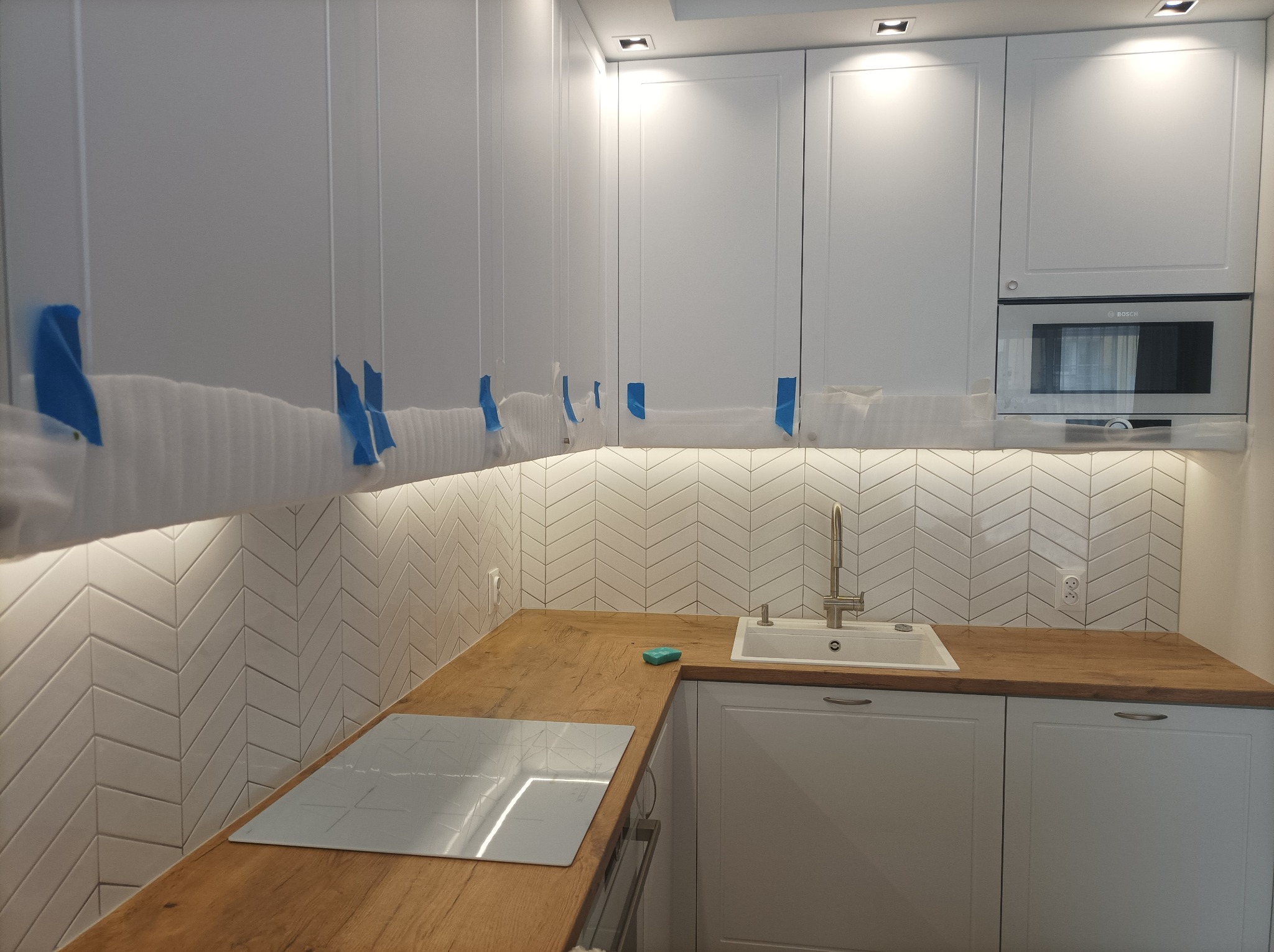 Nowoczesna kuchnia w trakcie wykończenia: białe szafki z drewnianym blatem, biała glazura ułożona w jodełkę, zabezpieczone folią ochronną górne szafki, widoczna płyta indukcyjna i zlewozmywak.