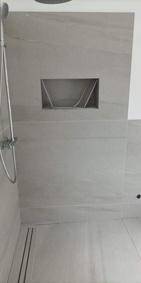 Wykończona ściana prysznicowa z szarych, matowych płytek imitujących kamień, wbudowana półka z ukrytymi przewodami, odpływ liniowy w podłodze.