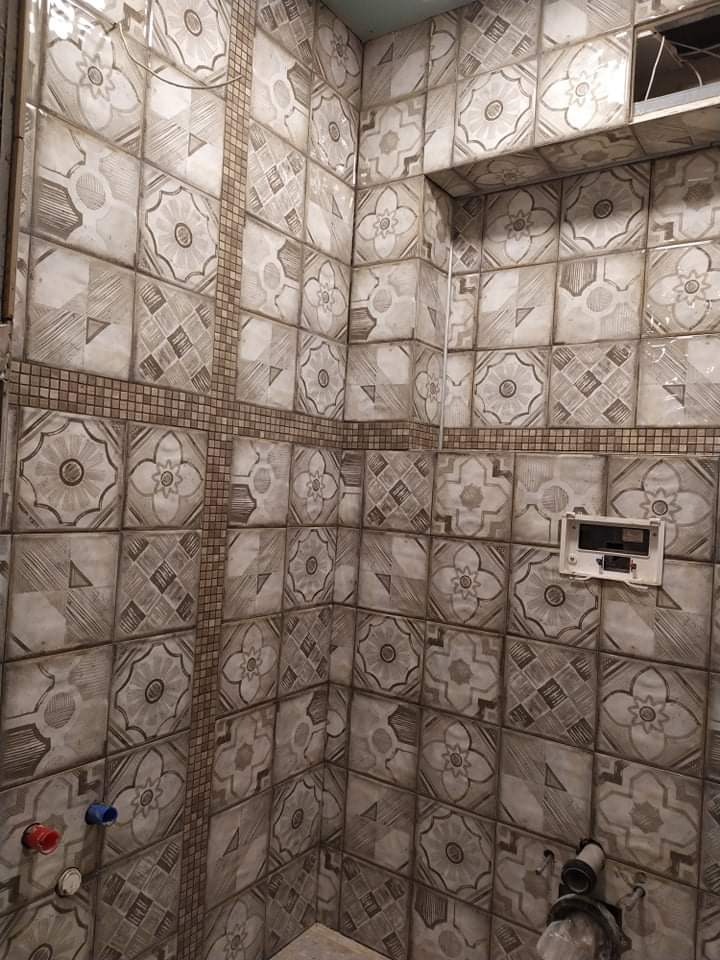 Wykończona ściana łazienki z kwadratowymi, wzorzystymi płytkami ceramicznymi w odcieniach szarości, z dekoracyjnymi pasami mozaiki i wystającymi rurami instalacji wodnej.