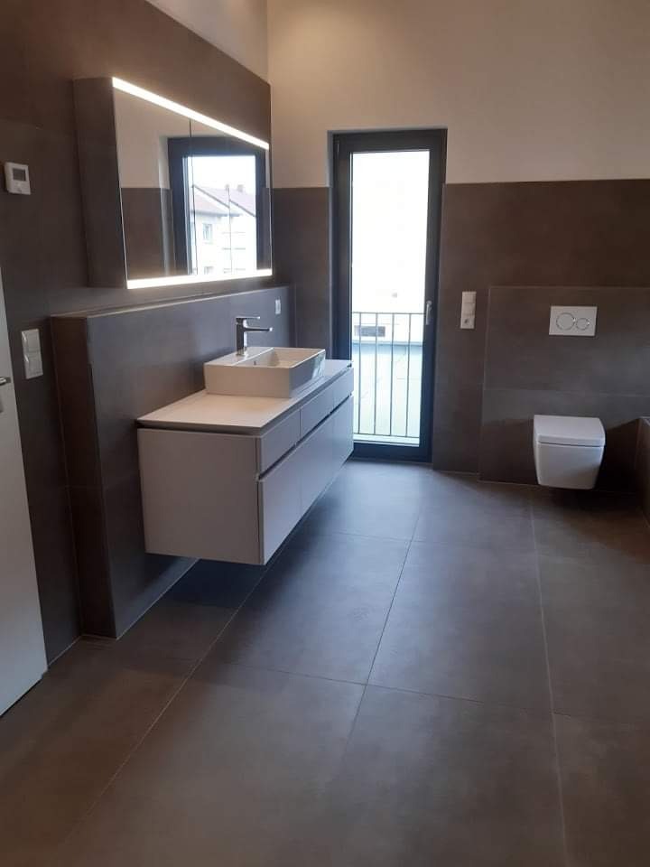 Nowoczesna łazienka z dużymi, szarymi płytkami podłogowymi i ściennymi, wisząca szafka pod umywalką z białą, prostokątną umywalką, podświetlane lustro i drzwi balkonowe z czarną ramą.