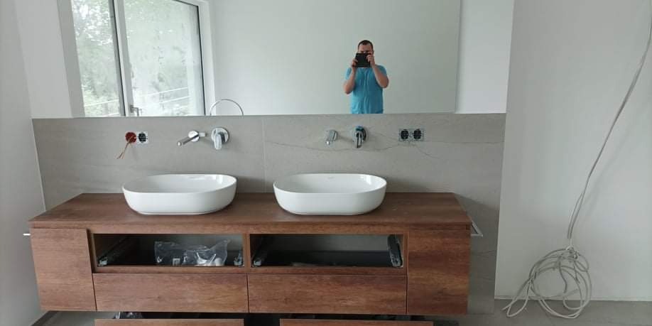 Dwie białe, owalne umywalki nablatowe na drewnianej szafce z otwartymi półkami i szufladami w trakcie montażu, widoczne niedokończone podłączenia elektryczne i hydrauliczne, odbicie fotografa...