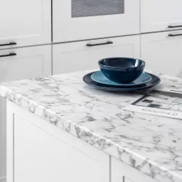 Biała kuchnia z marmurowym blatem, na którym leżą niebieskie naczynia i magazyn o kuchniach, fronty szafek z czarnymi uchwytami.