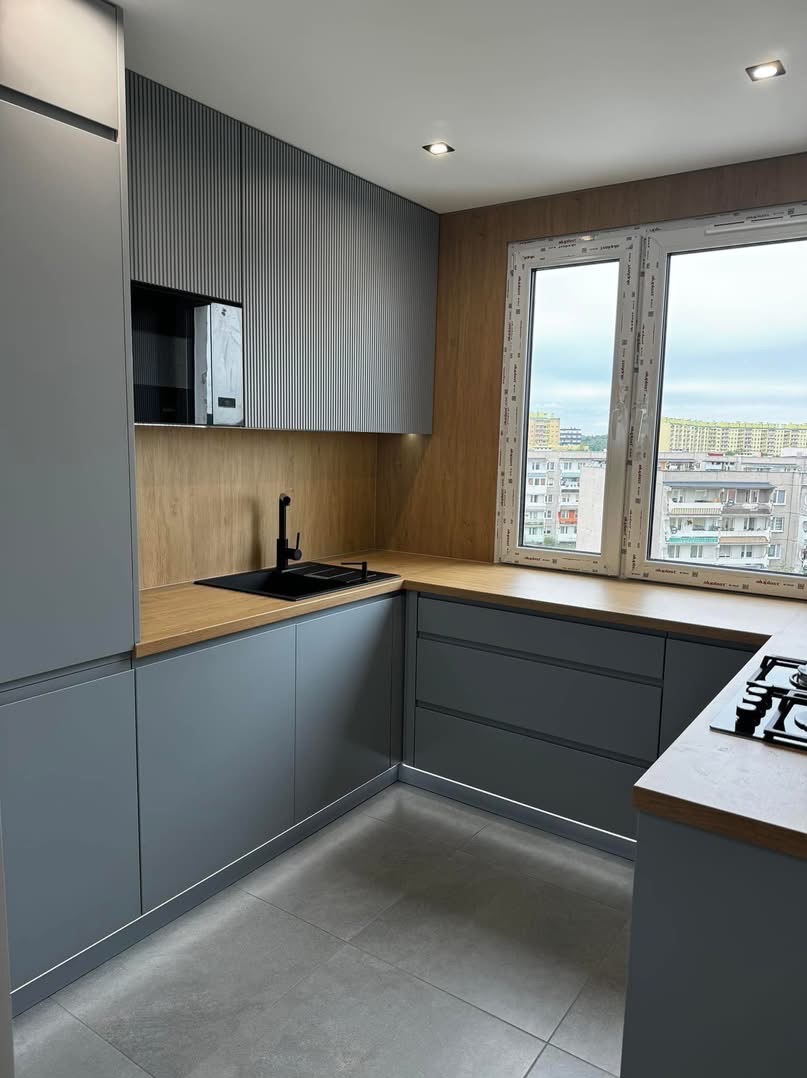 Szara kuchnia z drewnianym blatem i frontami z pionowymi żłobieniami, czarny zlew i bateria, okno z widokiem na miasto. Nowoczesny design.