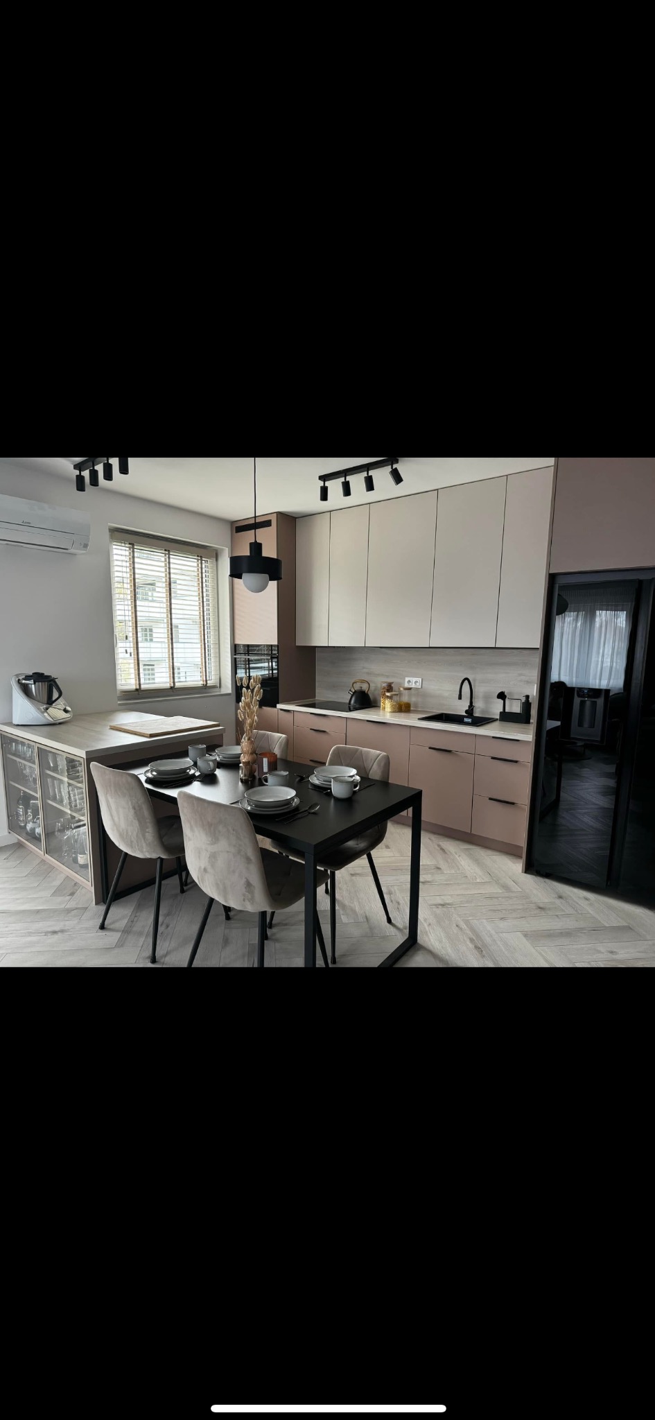 Stylowa kuchnia z wyspą, beżowymi szafkami i czarnymi akcentami oświetleniowymi. Na wyspie stół z nakryciem i krzesłami. Parkiet ułożony w jodełkę.