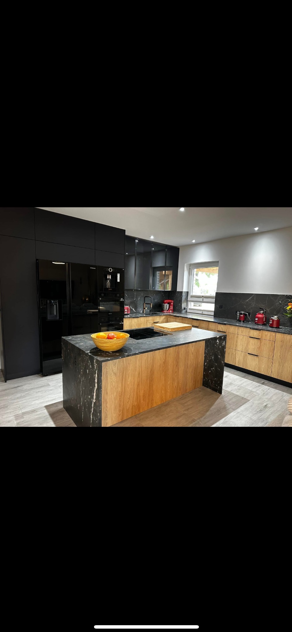 Nowoczesna kuchnia z wyspą: czarne szafki górne, dół w odcieniach drewna, marmurowy blat. Na wyspie misa z owocami, okno w tle. Minimalistyczny design.