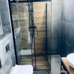 Nowoczesna łazienka z kabiną prysznicową typu walk-in z czarnymi profilami, drewnopodobnymi płytkami w strefie prysznica i szarymi płytkami na pozostałych ścianach, wisząca toaleta i umywalka...