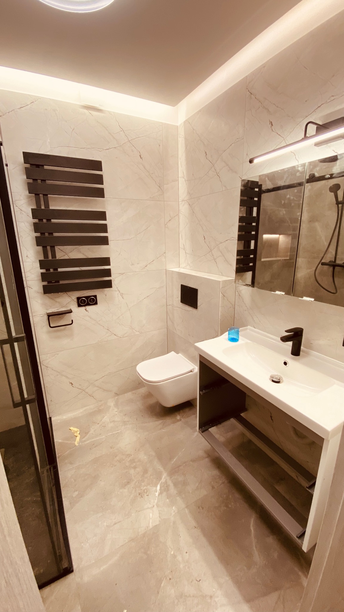 Wykończona łazienka z marmurowymi płytkami, wiszącą toaletą, grzejnikiem drabinkowym i umywalką z czarną armaturą. Widoczne detale wykończeniowe i oświetlenie.