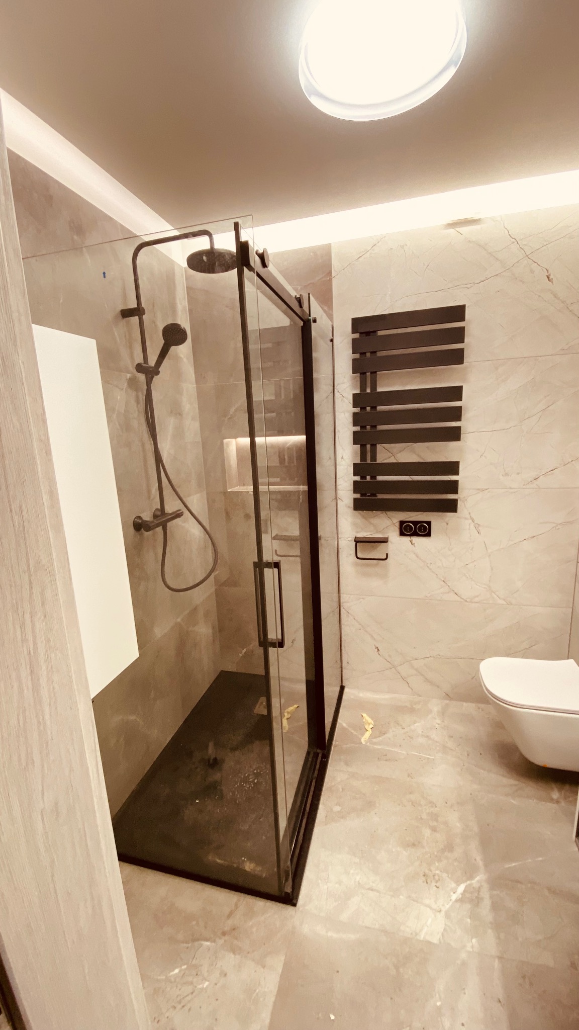 Wykończona łazienka z kabiną prysznicową typu walk-in z czarnymi ramami, marmurowymi płytkami i grzejnikiem drabinkowym. Nowoczesny design.