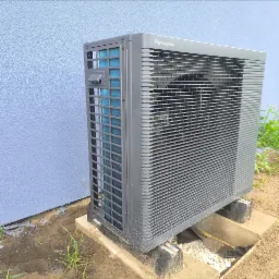 Szara pompa ciepła Panasonic Aquarea, ustawiona na betonowych blokach przy jasnoniebieskiej ścianie budynku. Widoczne detale konstrukcyjne i otoczenie.