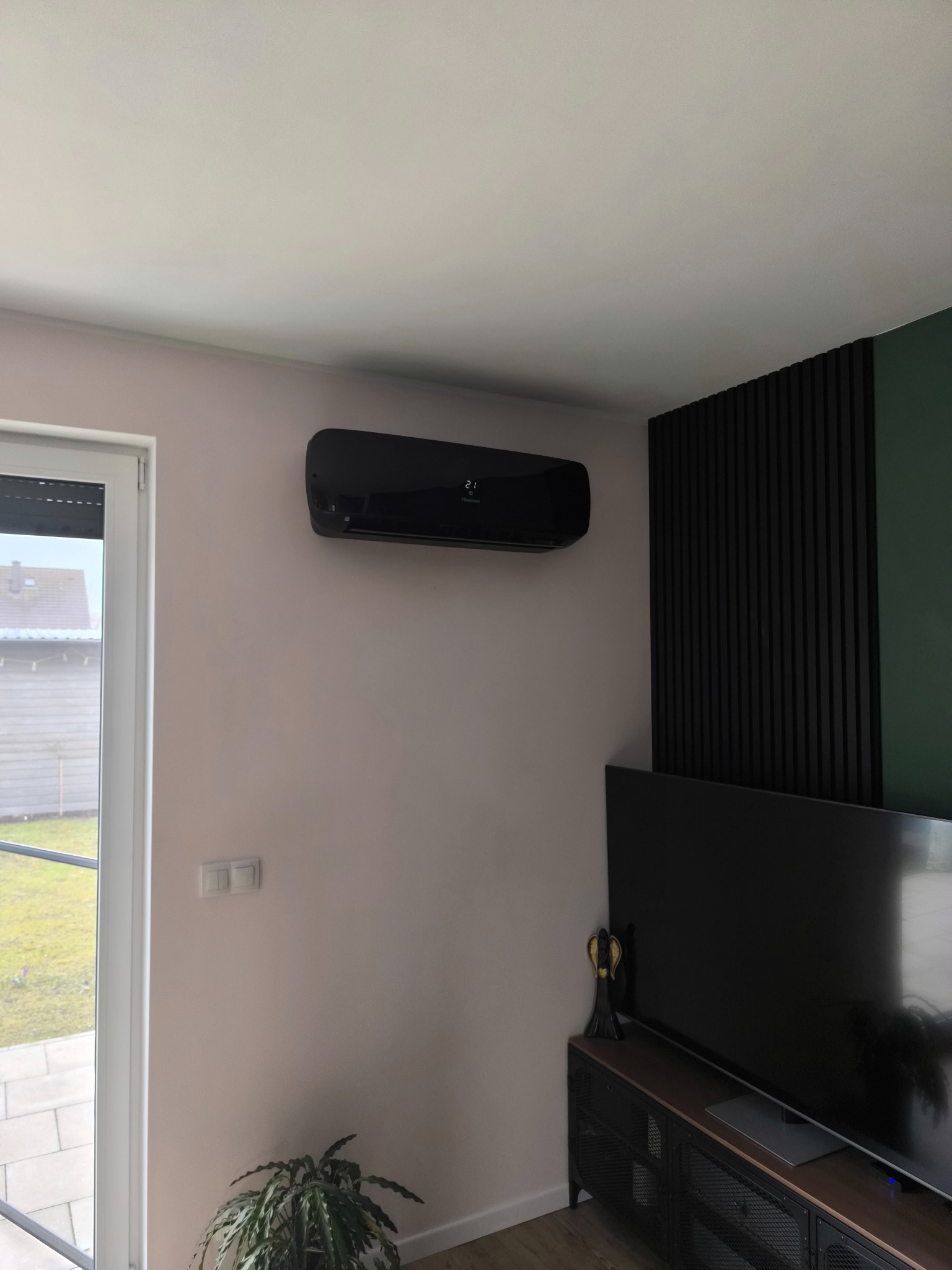Czarny klimatyzator ścienny Hisense zamontowany w nowoczesnym salonie z dekoracyjną ścianą z czarnych listew i zielonym akcentem, widok z dołu na urządzenie, obok okno z widokiem na ogród...