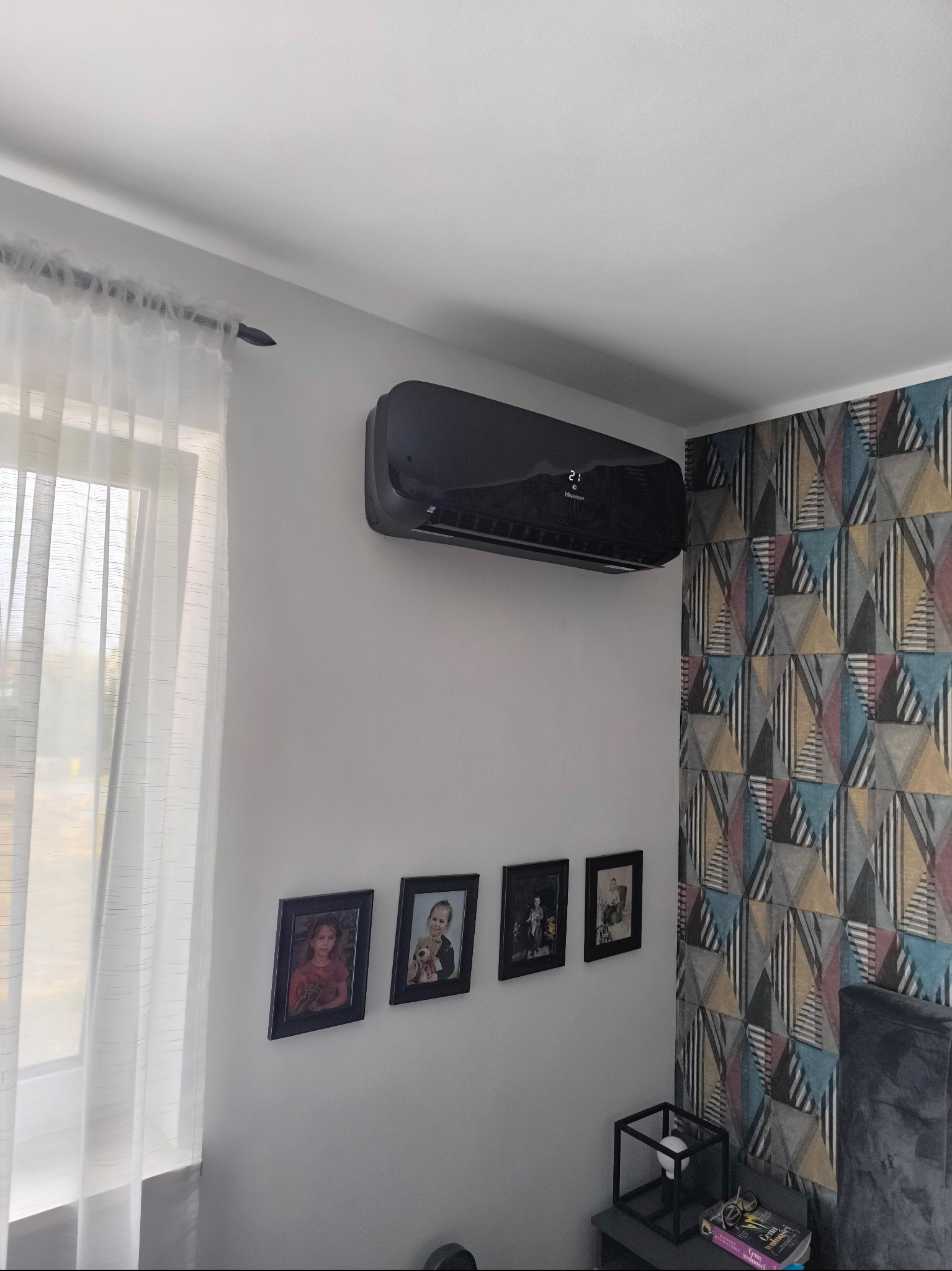 Czarny klimatyzator ścienny marki Hisense zamontowany w pokoju z wzorzystą tapetą, pod nim galeria czarno-białych zdjęć rodzinnych.