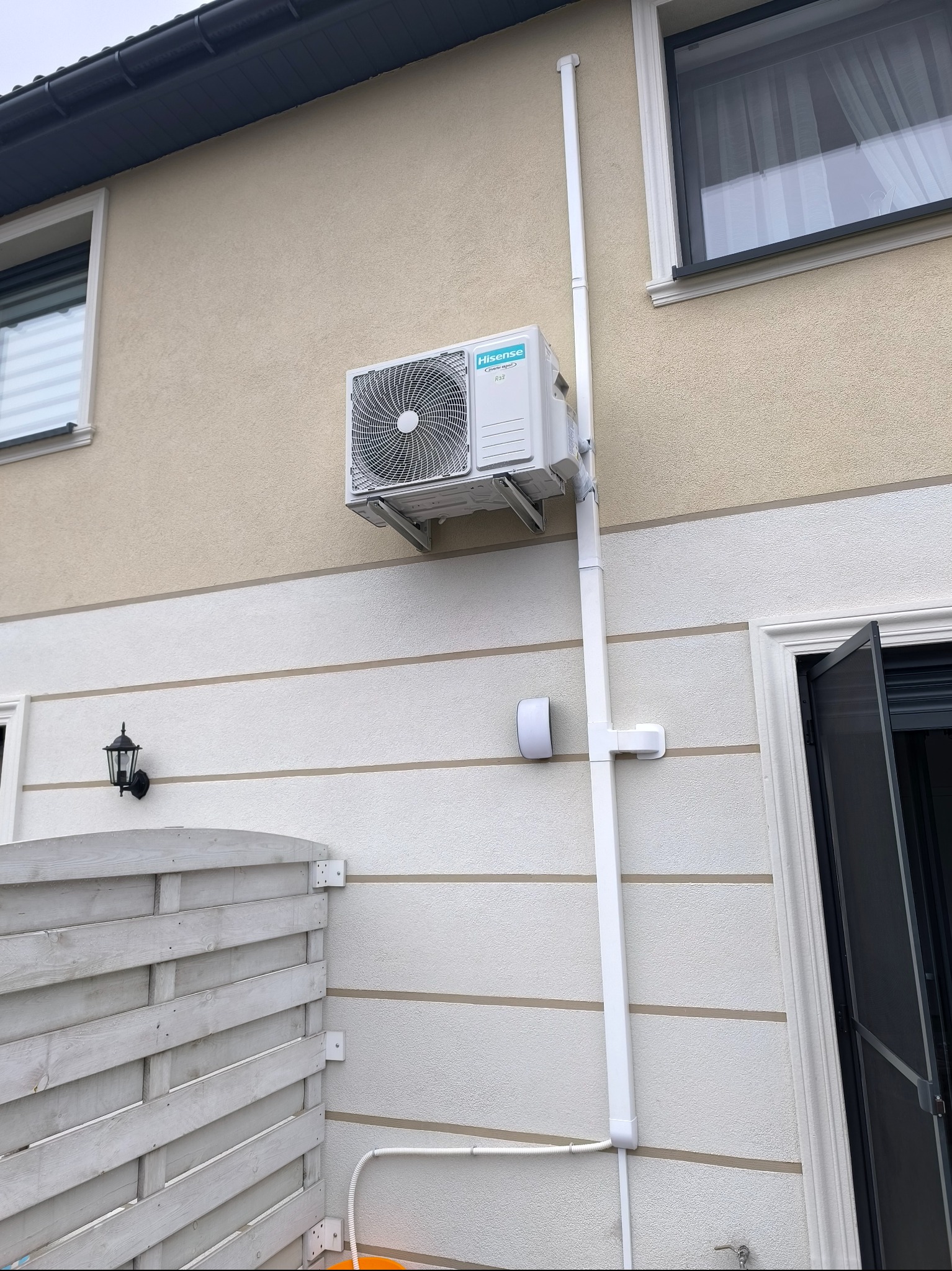 Zewnętrzna jednostka klimatyzacji Hisense zamontowana na elewacji domu z widocznymi rurami odprowadzającymi skropliny w białej osłonie.