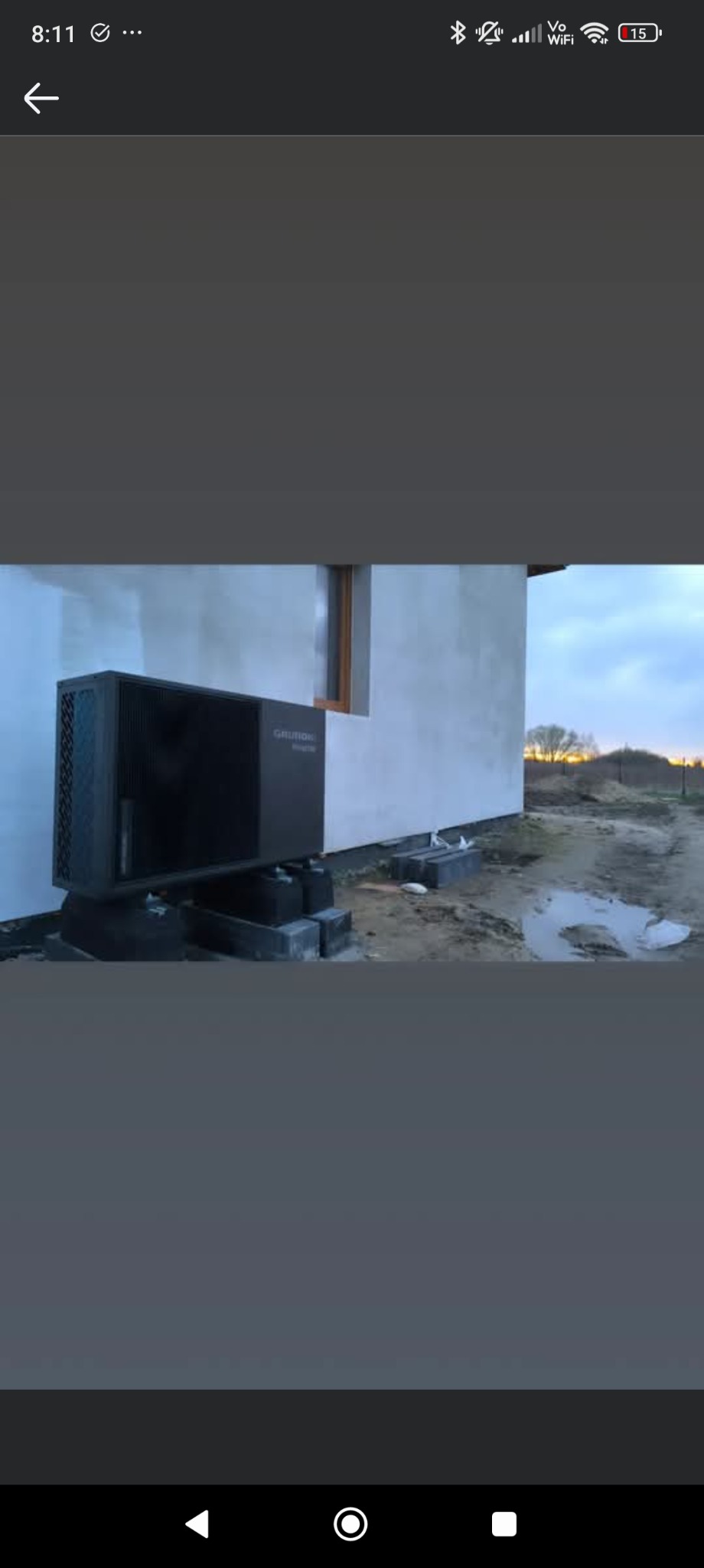 Zewnętrzna jednostka pompy ciepła marki Grundfos zainstalowana przy ścianie domu, na betonowych podstawach, widok na budynek z małym oknem i fragment krajobrazu w tle.