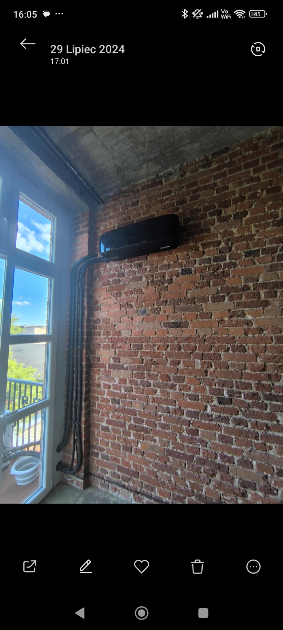 Czarny klimatyzator ścienny zamontowany na ceglanej ścianie w loftowym wnętrzu, widoczne rury instalacyjne prowadzone wzdłuż okna.