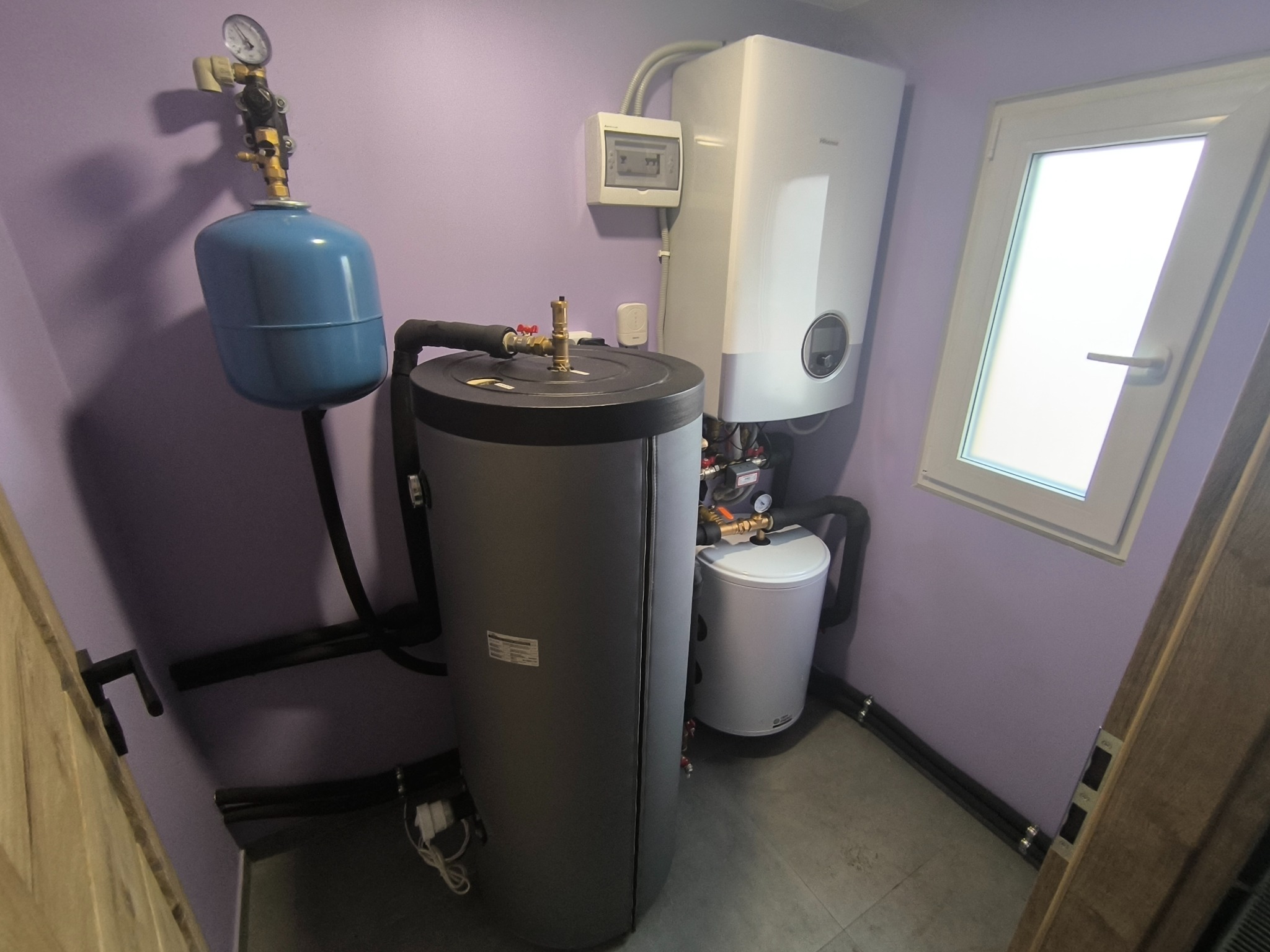 Kotłownia w pomieszczeniu z fioletowymi ścianami: zbiornik akumulacyjny, wiszący kocioł grzewczy oraz naczynie przeponowe z manometrem.