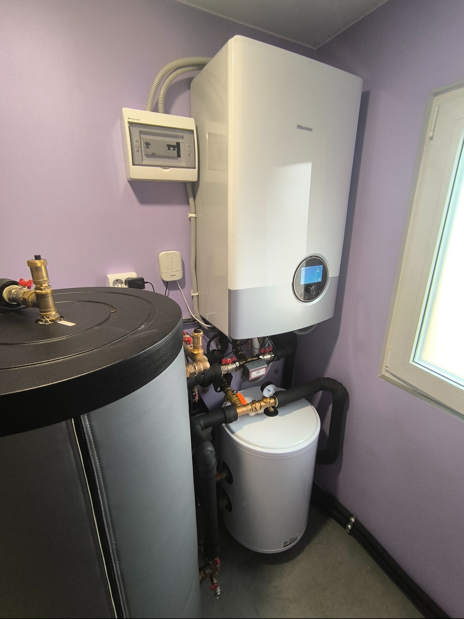 Nowoczesna instalacja grzewcza z zbiornikiem buforowym, pompą ciepła Hisense i rozdzielnicą elektryczną na fioletowej ścianie w pomieszczeniu z oknem.