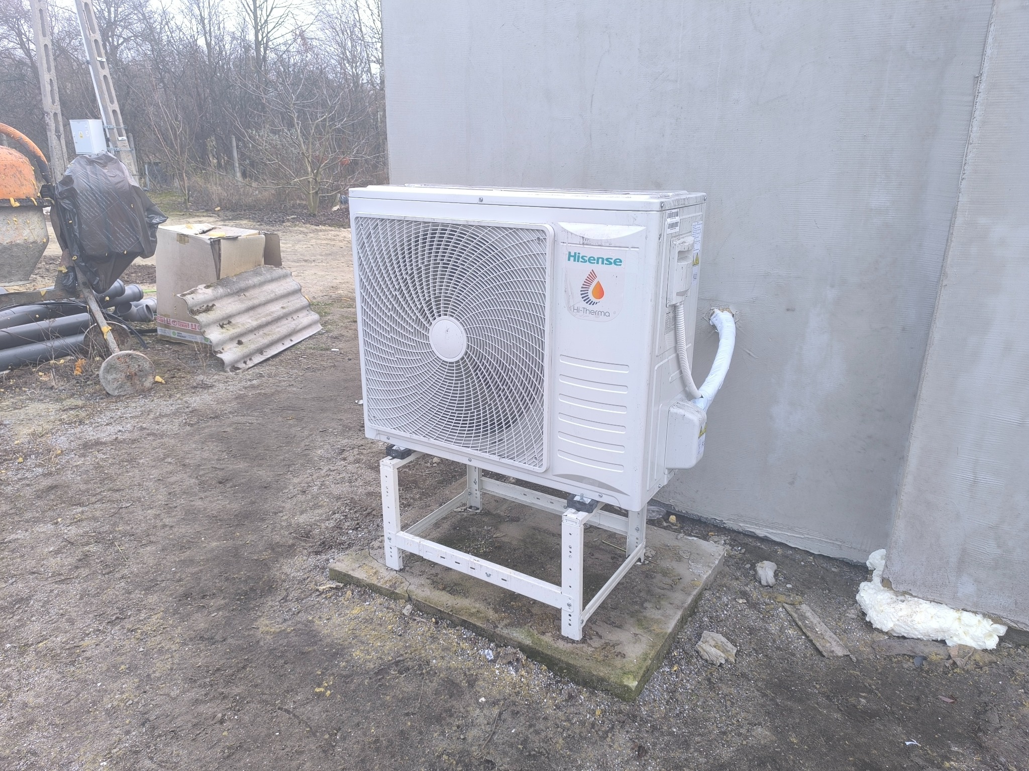 Biała zewnętrzna jednostka pompy ciepła marki Hisense, zamontowana na metalowym stelażu na betonowej płycie, przy szarej ścianie budynku. Widoczne elementy instalacji i pianka montażowa przy ścianie.
