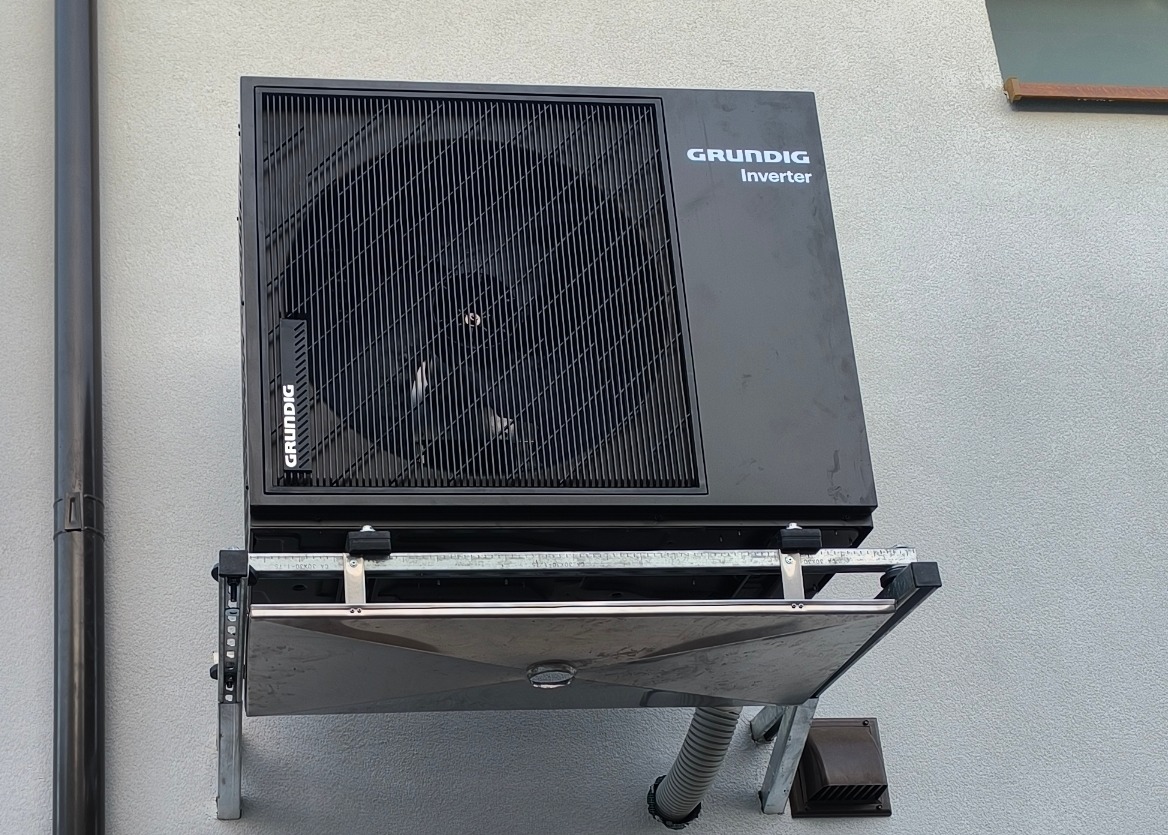 Czarna pompa ciepła Grundig Inverter zamontowana na zewnątrz budynku na metalowym stelażu z otwartą dolną osłoną. Widoczny szary przewód odprowadzający skropliny.