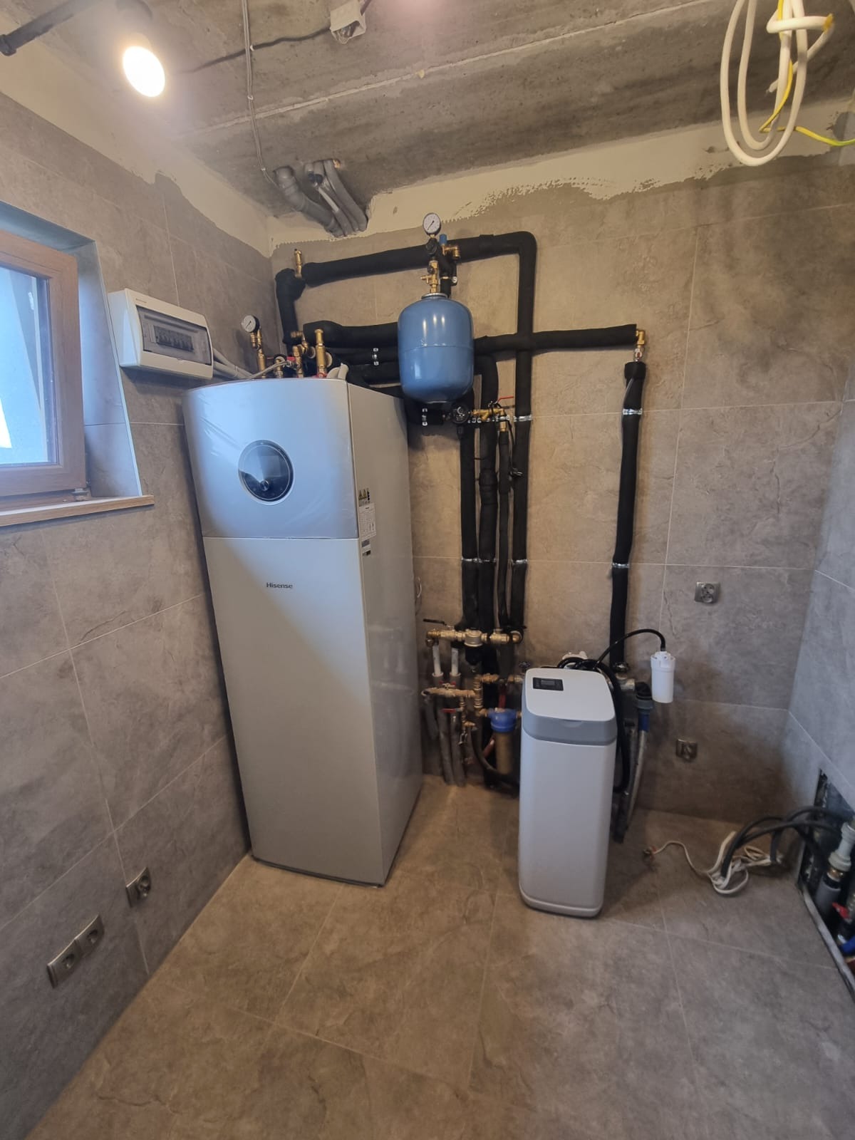 Nowoczesna pompa ciepła Hisense i instalacja w pomieszczeniu kotłowni w Ratowicach. Widoczne rury, zawory i zbiornik wyrównawczy. Wykończenie w płytkach.