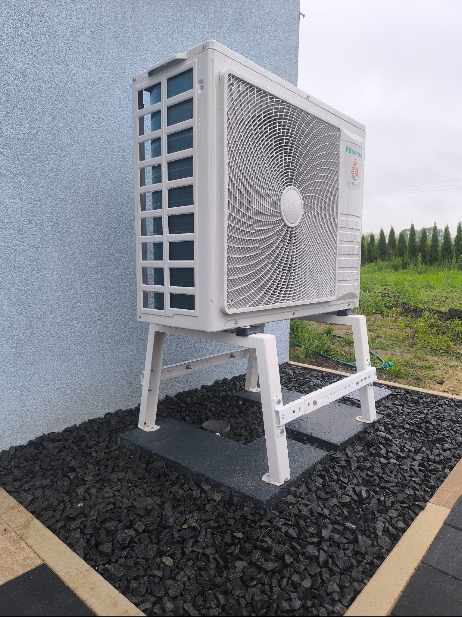 Biała pompa ciepła Hisense na wsporniku, zainstalowana przy ścianie budynku, otoczona czarnym grysem. Widok na zewnątrz, zieleń w tle.