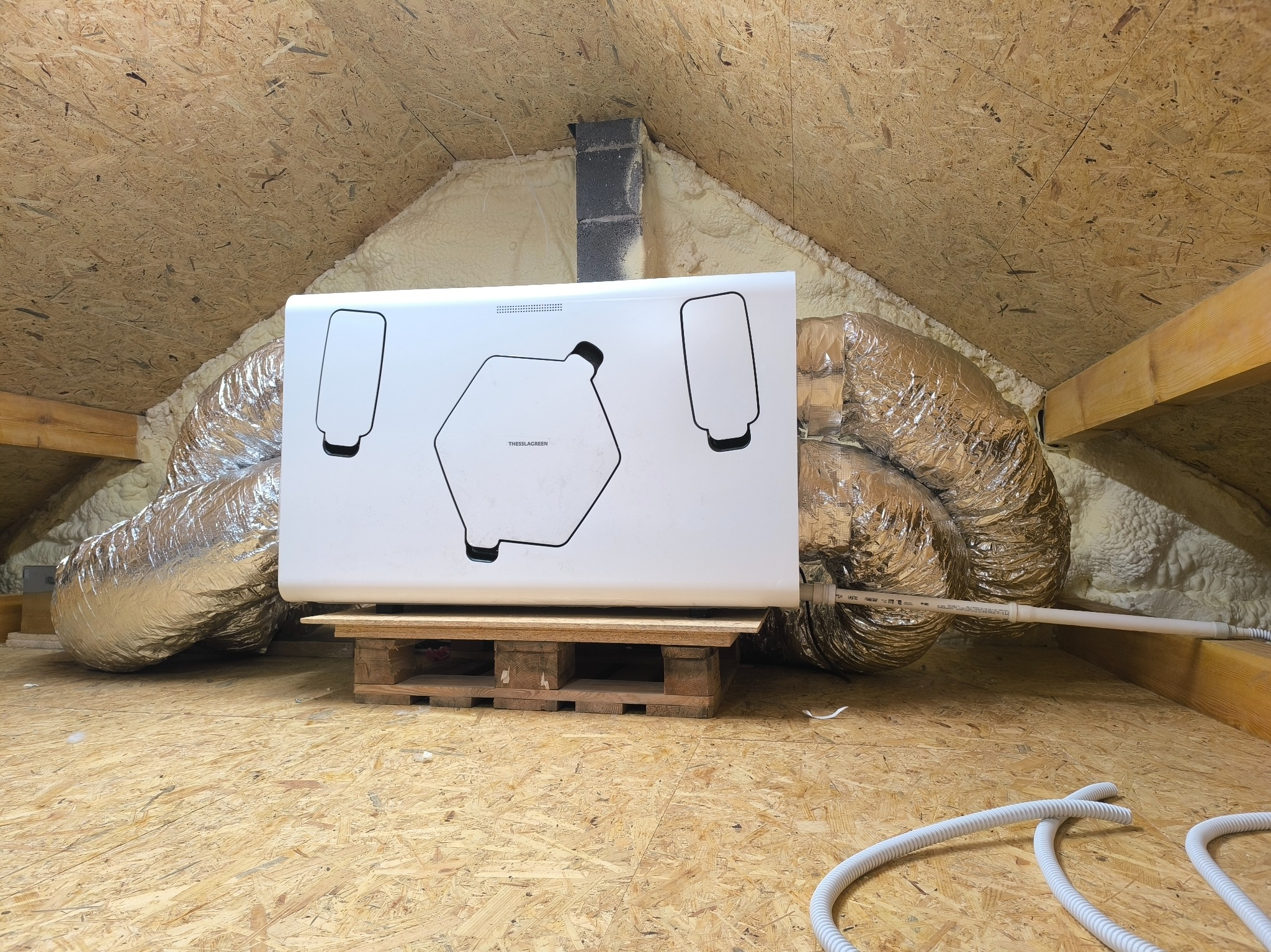 Centrala wentylacyjna z rekuperacją na poddaszu, widoczne przewody wentylacyjne i izolacja piankowa. Urządzenie stoi na drewnianej palecie.