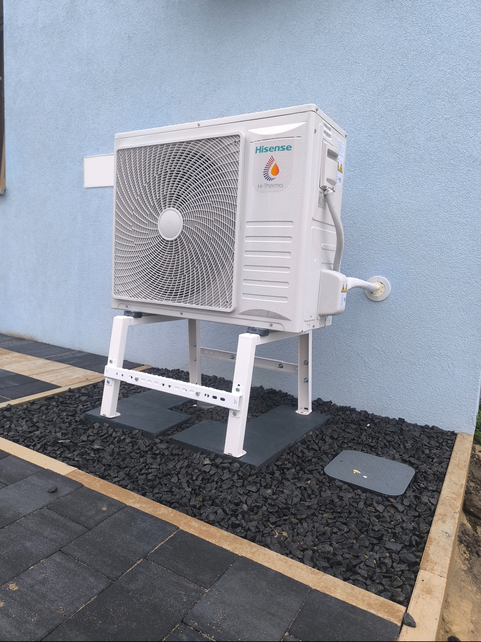Biała pompa ciepła Hisense na metalowym stelażu, zamontowana przy jasnoniebieskiej ścianie budynku. Podłoże wyłożone czarnym kamieniem i szarymi płytami.