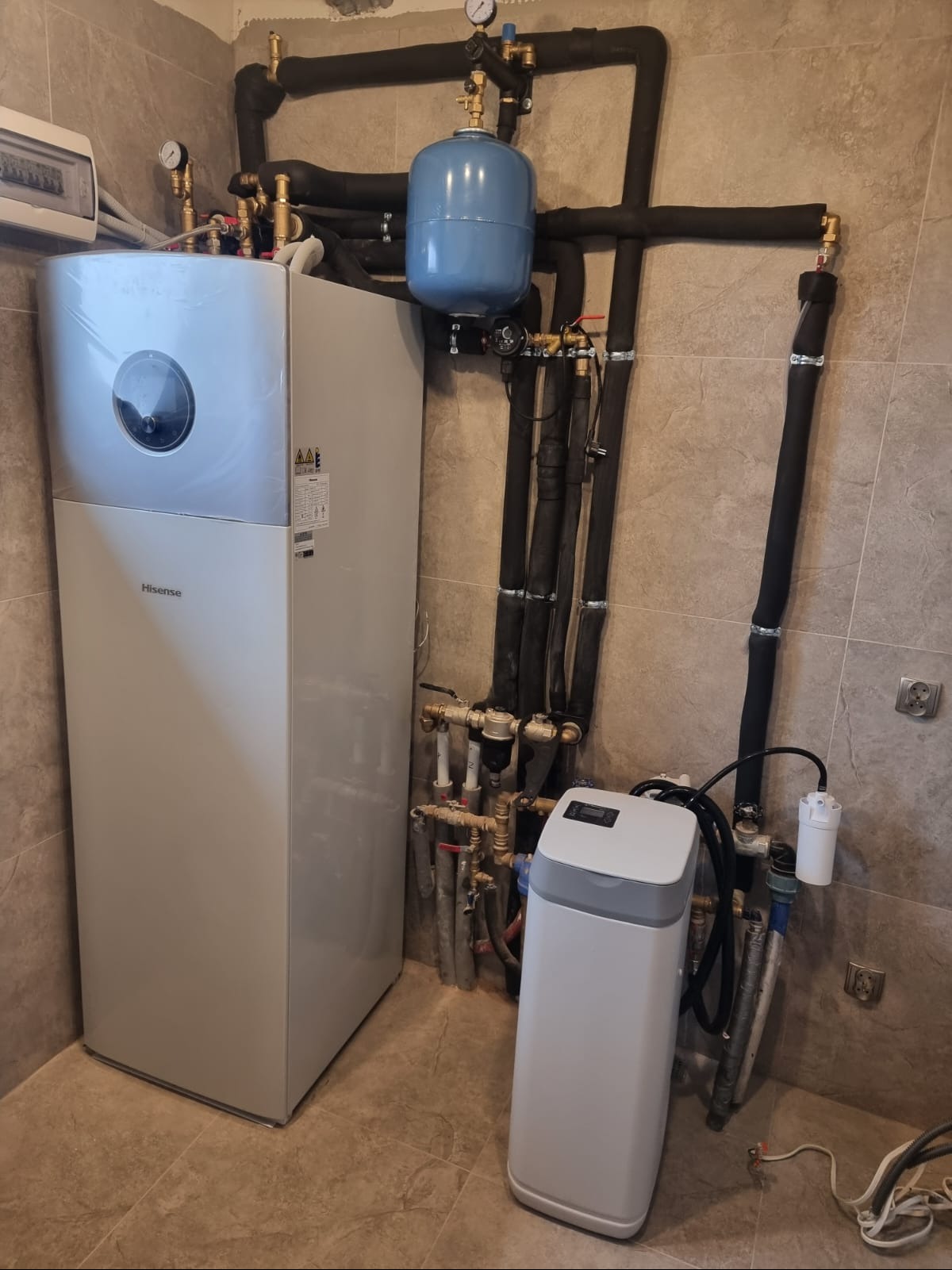 Nowoczesna instalacja pompy ciepła Hisense z zbiornikiem wyrównawczym i filtrem wody, rury w czarnej izolacji, manometry, w pomieszczeniu z beżowymi płytkami.