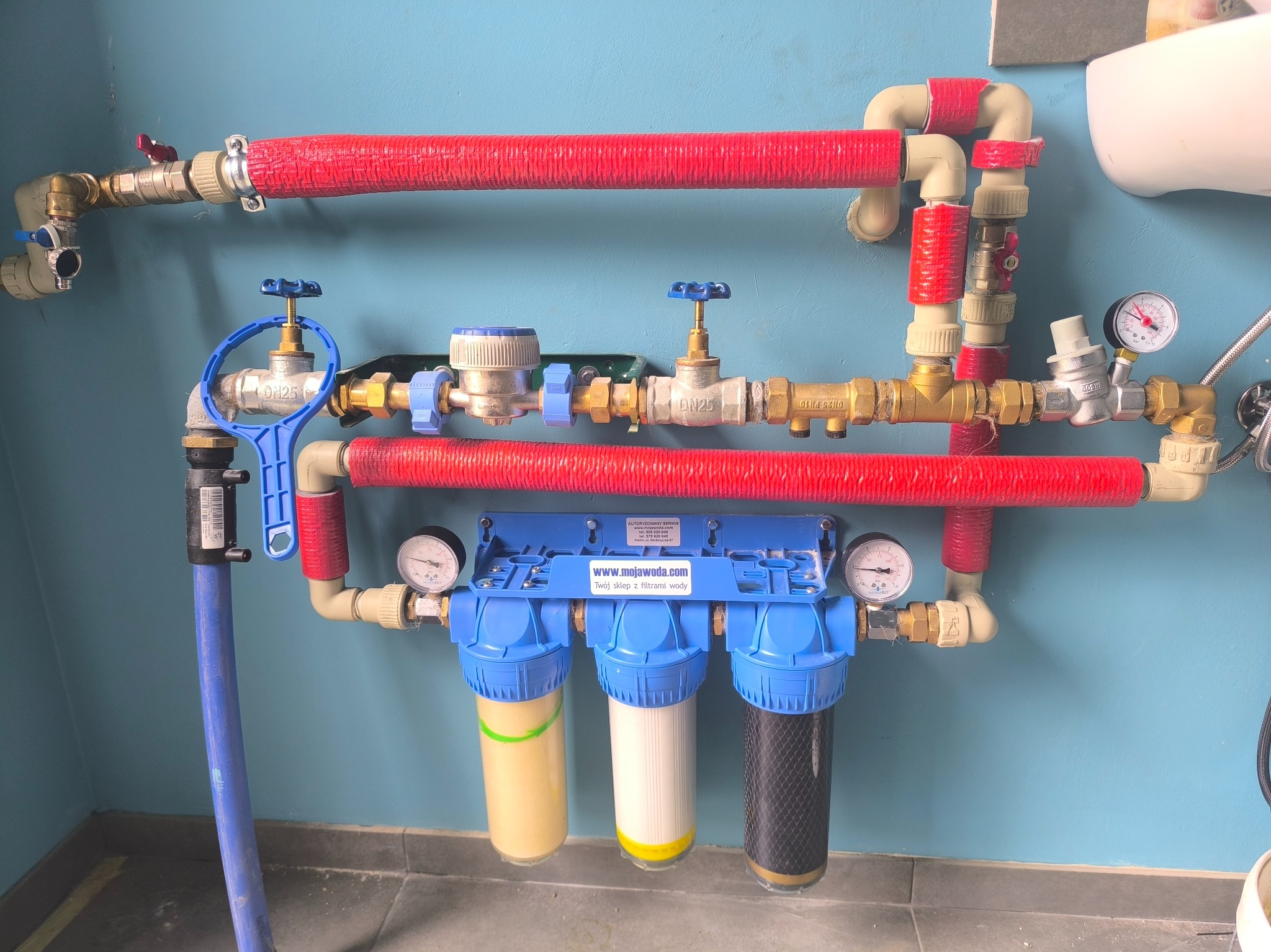 Instalacja filtrów wody z trzema cylindrycznymi filtrami (żółty, biały, czarny) i rurami owiniętymi czerwoną taśmą, zamontowana na błękitnej ścianie w pomieszczeniu.