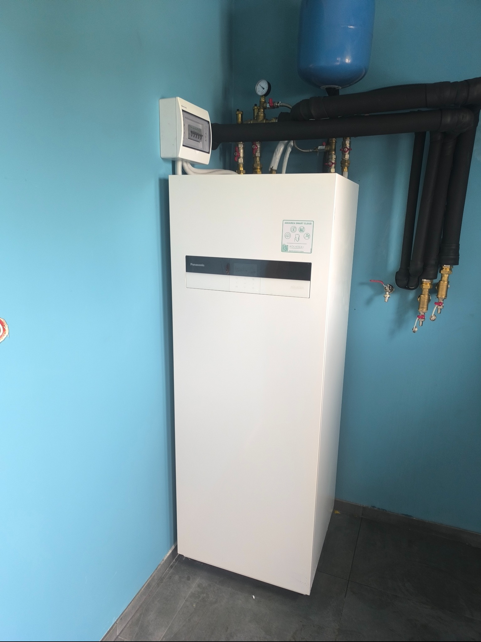 Biała pompa ciepła Panasonic Aquarea Smart Cloud, z czarnymi rurami i elementami instalacji, na tle turkusowej ściany i szarej podłogi. Widoczny licznik elektryczny.