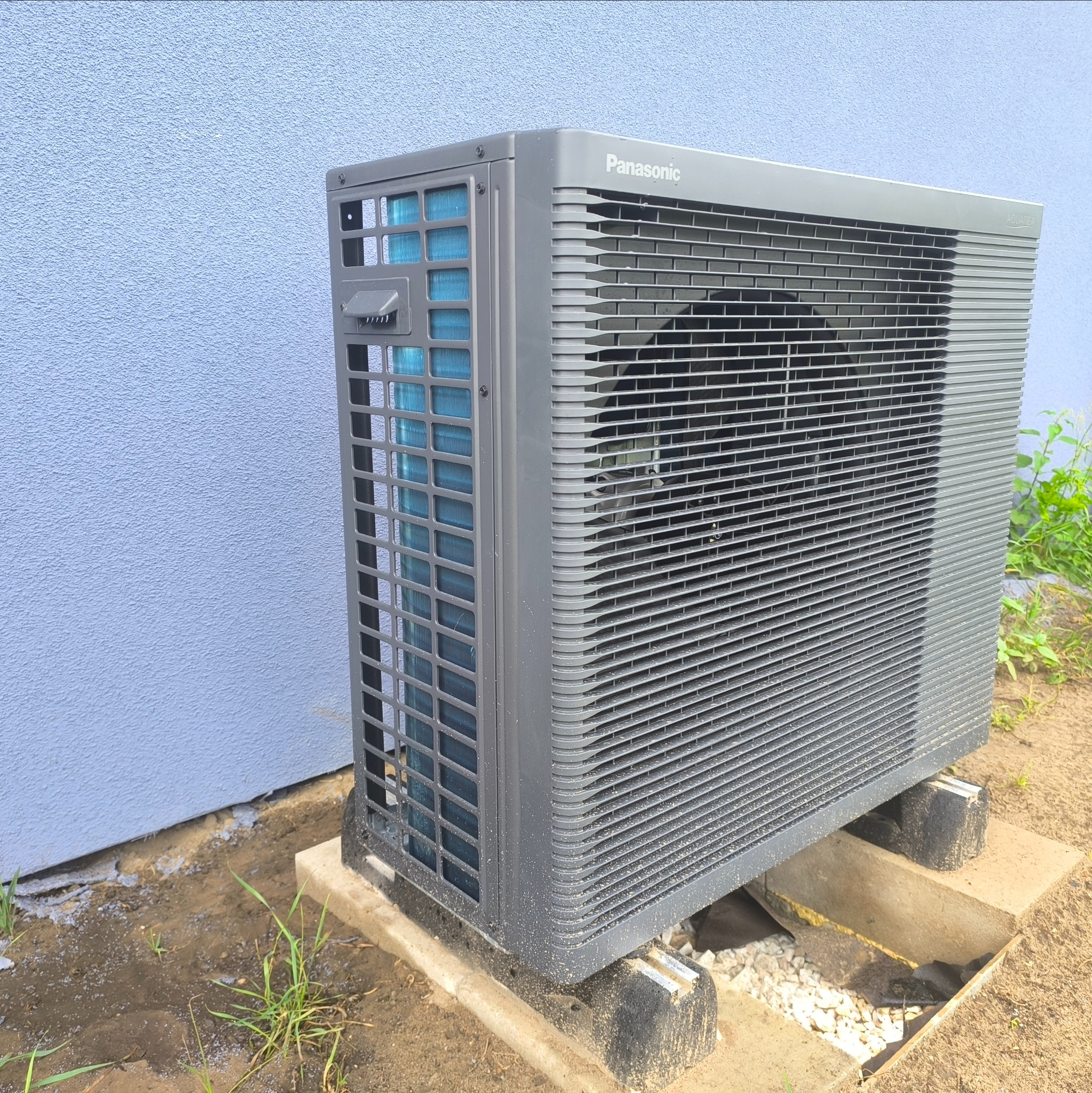 Szara pompa ciepła Panasonic Aquarea, ustawiona na betonowych blokach przy jasnoniebieskiej ścianie budynku. Widoczne detale konstrukcyjne i otoczenie.
