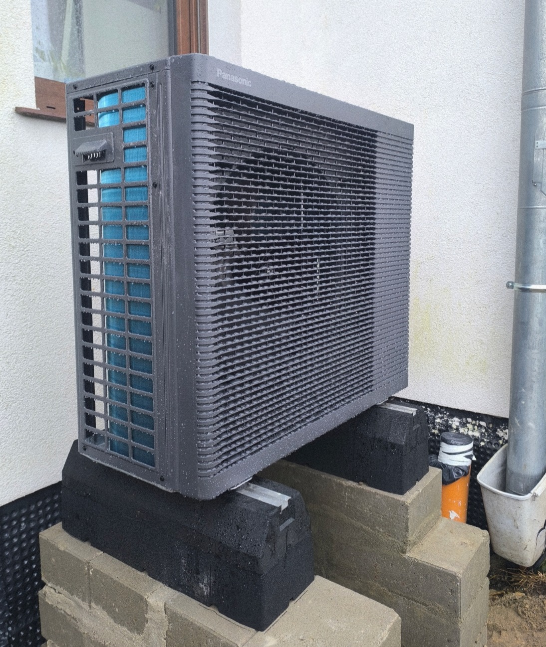 Szara pompa ciepła Panasonic Aquarea, zamontowana na betonowych blokach przy ścianie budynku. Widoczny fragment okna i rury spustowej. Krople wody na obudowie.