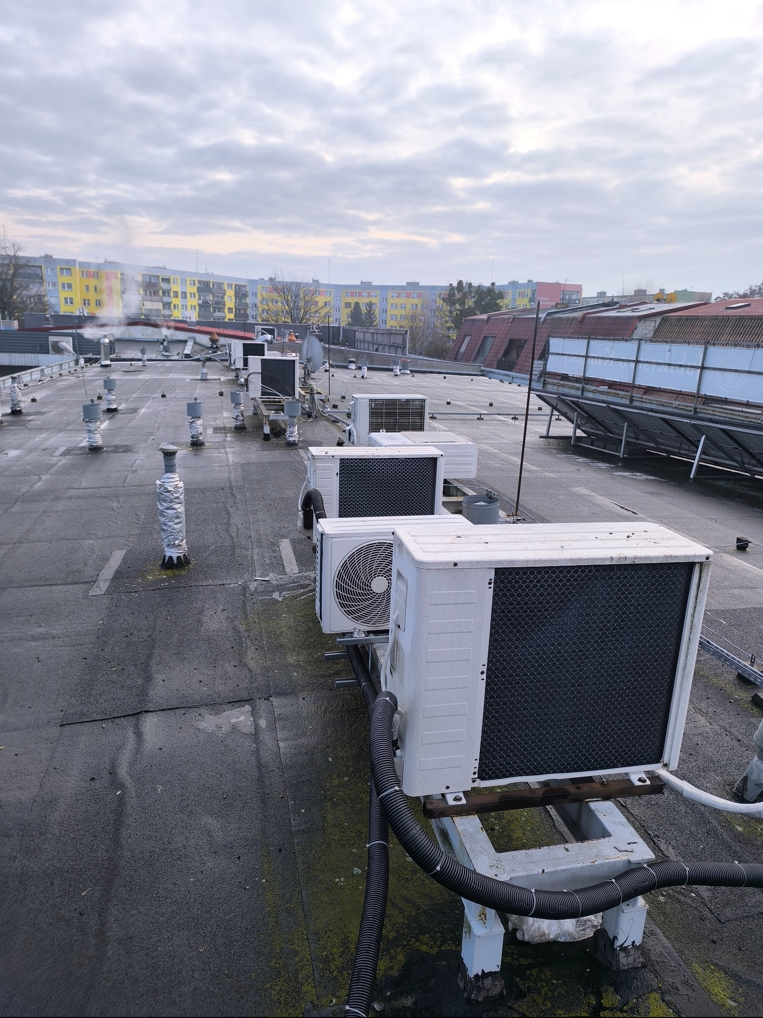 Rząd białych klimatyzatorów na płaskim dachu budynku, widok z góry, w tle bloki mieszkalne i pochmurne niebo. Widoczne rury i elementy montażowe.