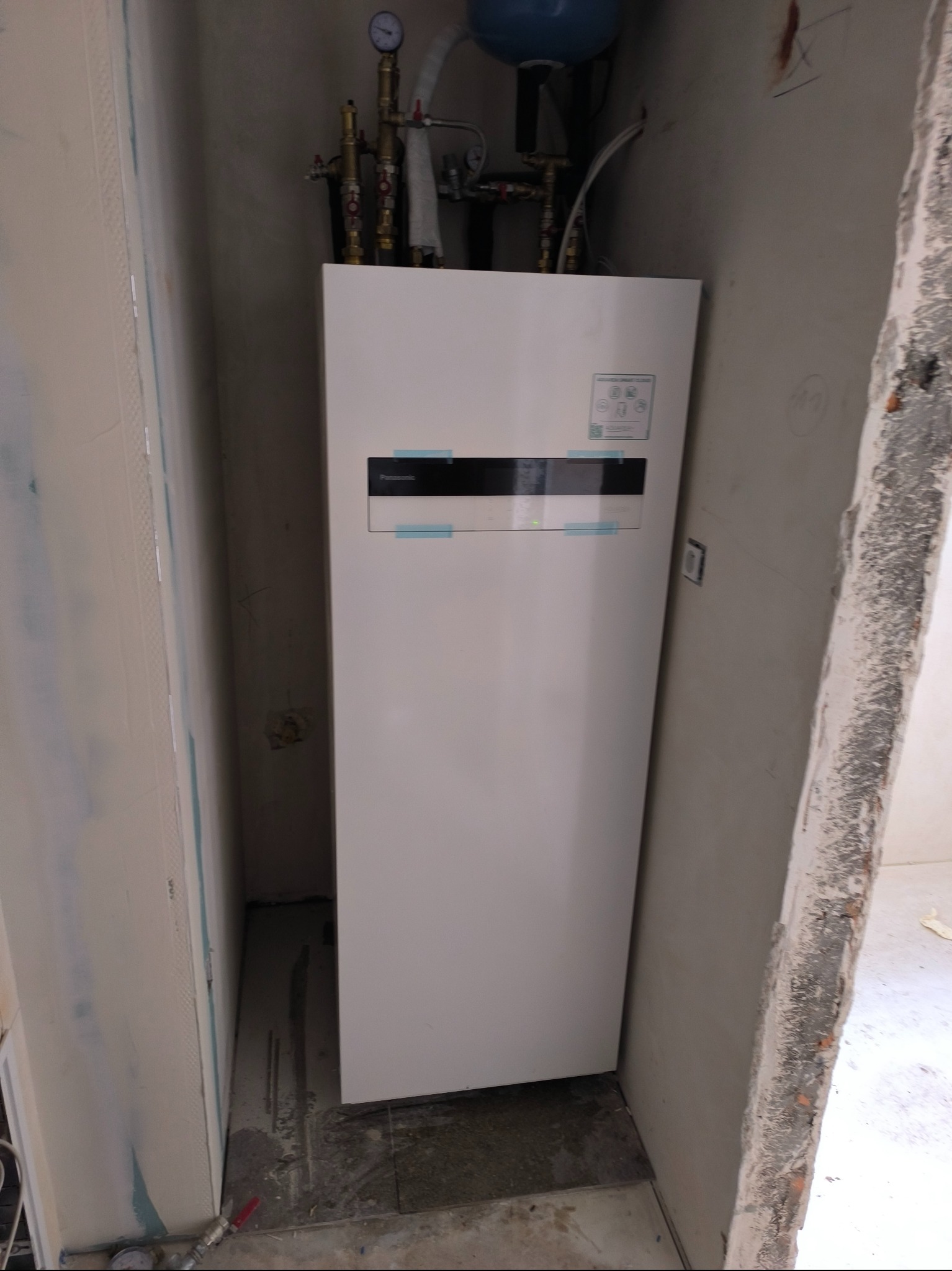 Biała pompa ciepła Panasonic Aquarea Smart Cloud, widoczna w rogu pomieszczenia w trakcie remontu. Na górze instalacja hydrauliczna i zbiornik wyrównawczy.
