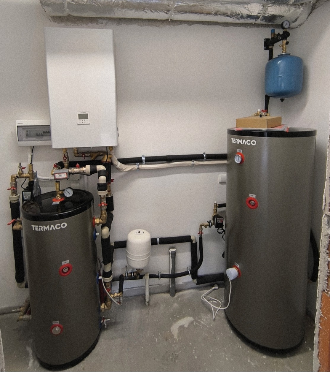Kompletna instalacja grzewcza z dwoma zbiornikami Termaco, białym piecem i niebieskim zbiornikiem wyrównawczym. Rury i zawory tworzą skomplikowany system.