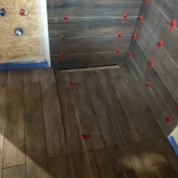 Układanie płytek imitujących drewno w narożniku pomieszczenia, z użyciem poziomych listew dystansowych i czerwonych klipsów poziomujących.