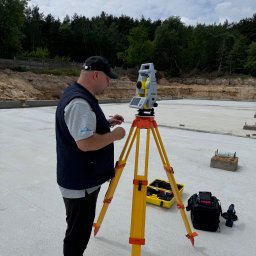 Usługi Geodezyjno-Kartograficzne BM-GEO Bogdan Stasiurka - Pracownik w czarnej kamizelce i czerwonych butach Nike obsługuje niwelator na statywie na wylanej płycie fundamentowej, w tle las i pochmurne niebo.