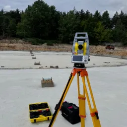 Tachimetr GeoMax Zenith 35 Pro na żółtym statywie, skrzynka narzędziowa i torba obok, widok na przygotowany fundament pod halę i las w tle, Okmiany.