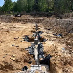 Wykonane fundamenty pod halę w Okmianach, widoczne betonowe słupy i czarna folia ochronna w wykopach, w tle drzewa i niebieskie niebo.