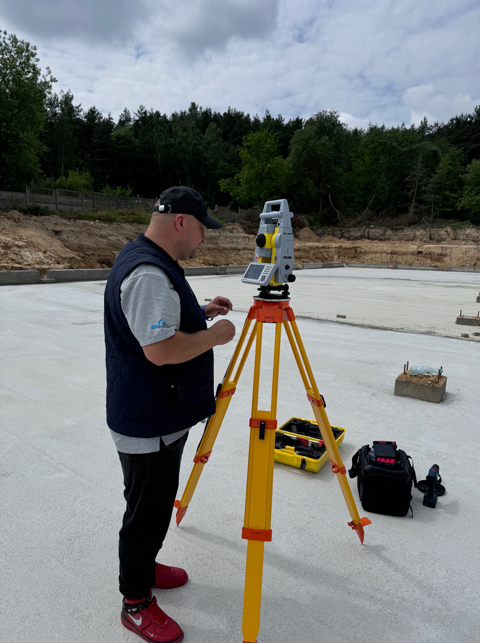 Pracownik w czarnej kamizelce i czerwonych butach Nike obsługuje niwelator na statywie na wylanej płycie fundamentowej, w tle las i pochmurne niebo.