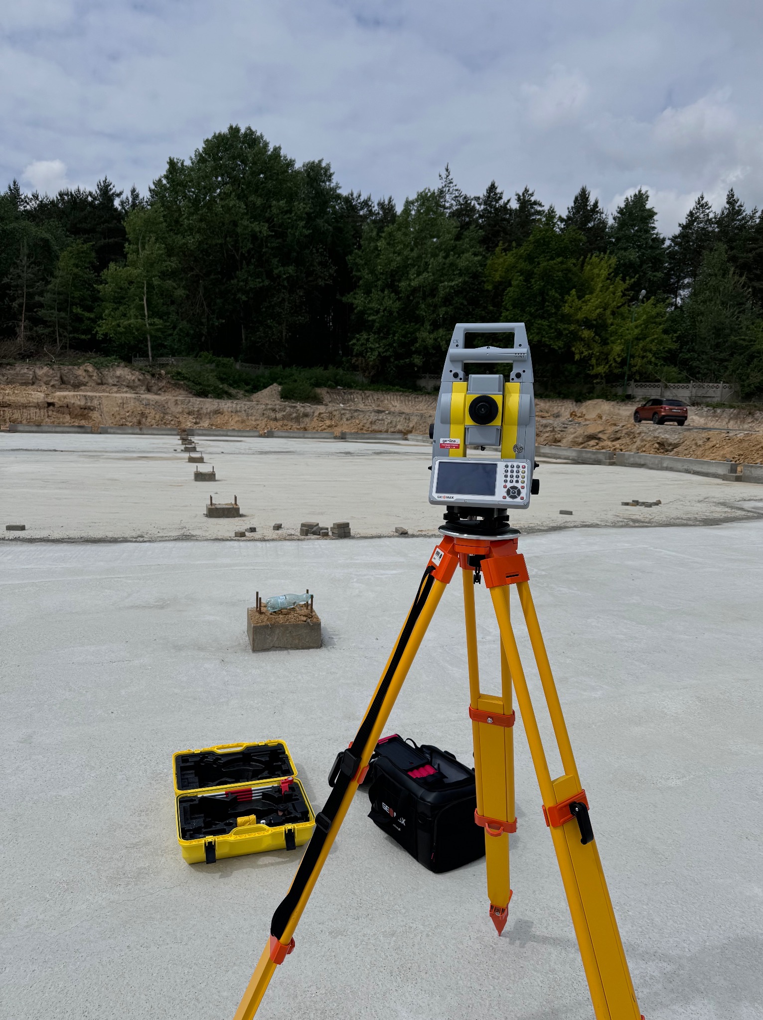 Tachimetr GeoMax Zenith 35 Pro na żółtym statywie, skrzynka narzędziowa i torba obok, widok na przygotowany fundament pod halę i las w tle, Okmiany.