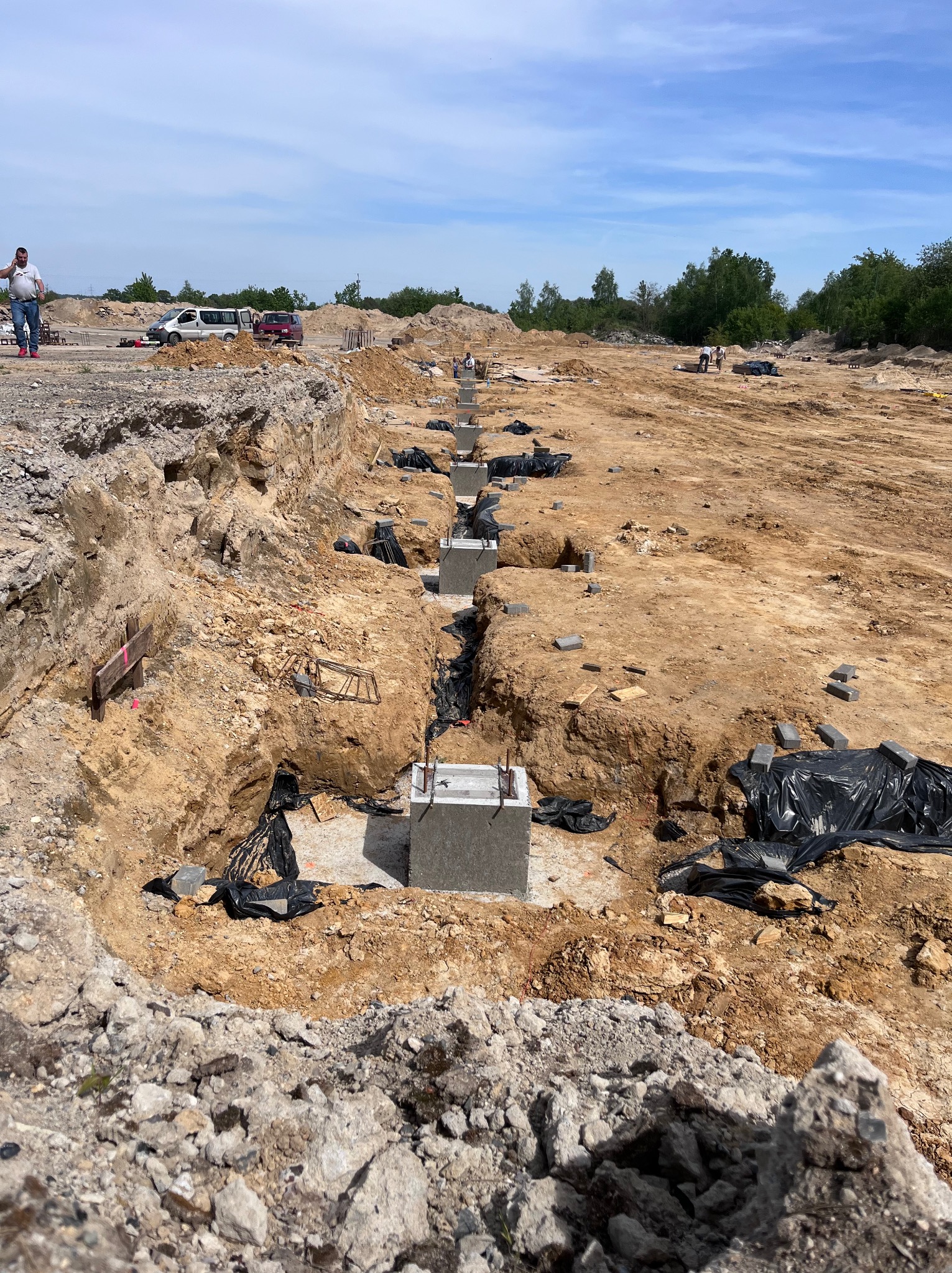 Wykopy pod fundamenty hali w Okmianach, widoczne betonowe podstawy z wystającymi prętami zbrojeniowymi, zabezpieczone czarną folią, w tle pracownicy i pojazdy na placu budowy.